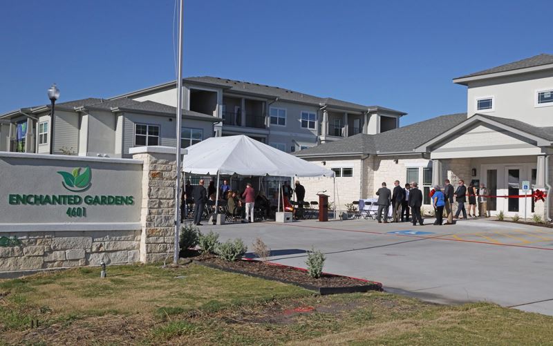 The front parking lot of an apartment complex. People are gathered under a white canopy.