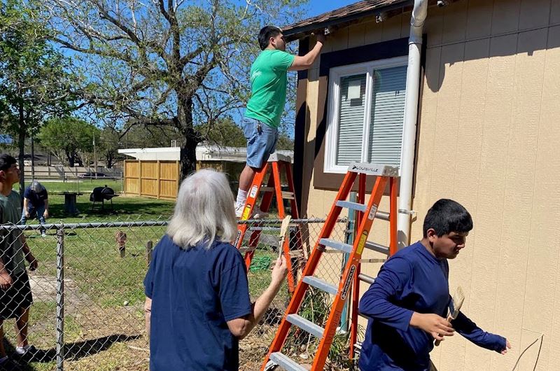 People help to apply a new coat of beige paint to a home. One person on ladder, one with a brush