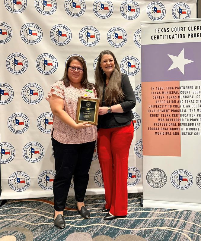 A woman accepts an award plaque from another woman in front of a TCCA backdrop