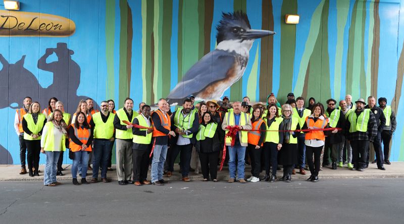Large group of people in safety vests cutting a ribbon in front of a mural featuring a kingfisher