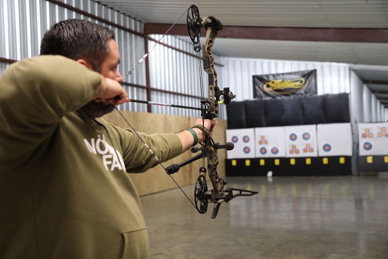 A man drawsback a bowstring to shoot a target indoors.