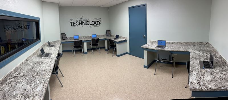 A computer lab with laptops set up on counters facing the wall. A logo on the wall reads Technology.