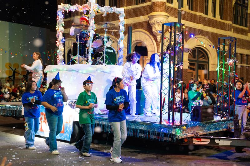 Parade float with a drummer on a stage. Kids walk alongside with Happy Birthday Jesus shirts.
