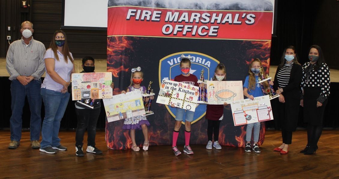 Children hold homemade posters on a stage. They are flanked by adults.