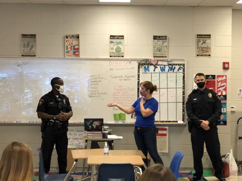 A man, a woman and another man stand in a row in front of a class. Woman talks to man on her right.