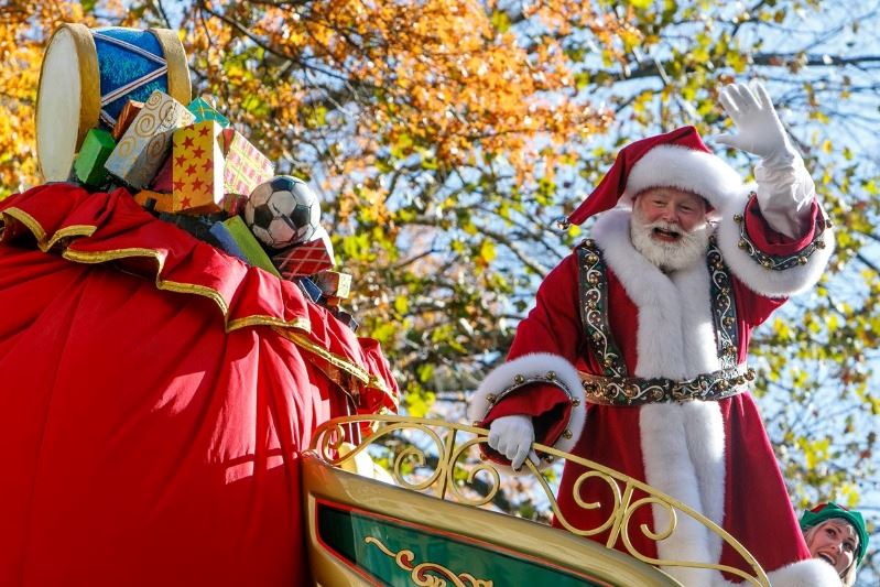Santa Claus waves from sleigh-themed parade float. Trees are visible in the background.