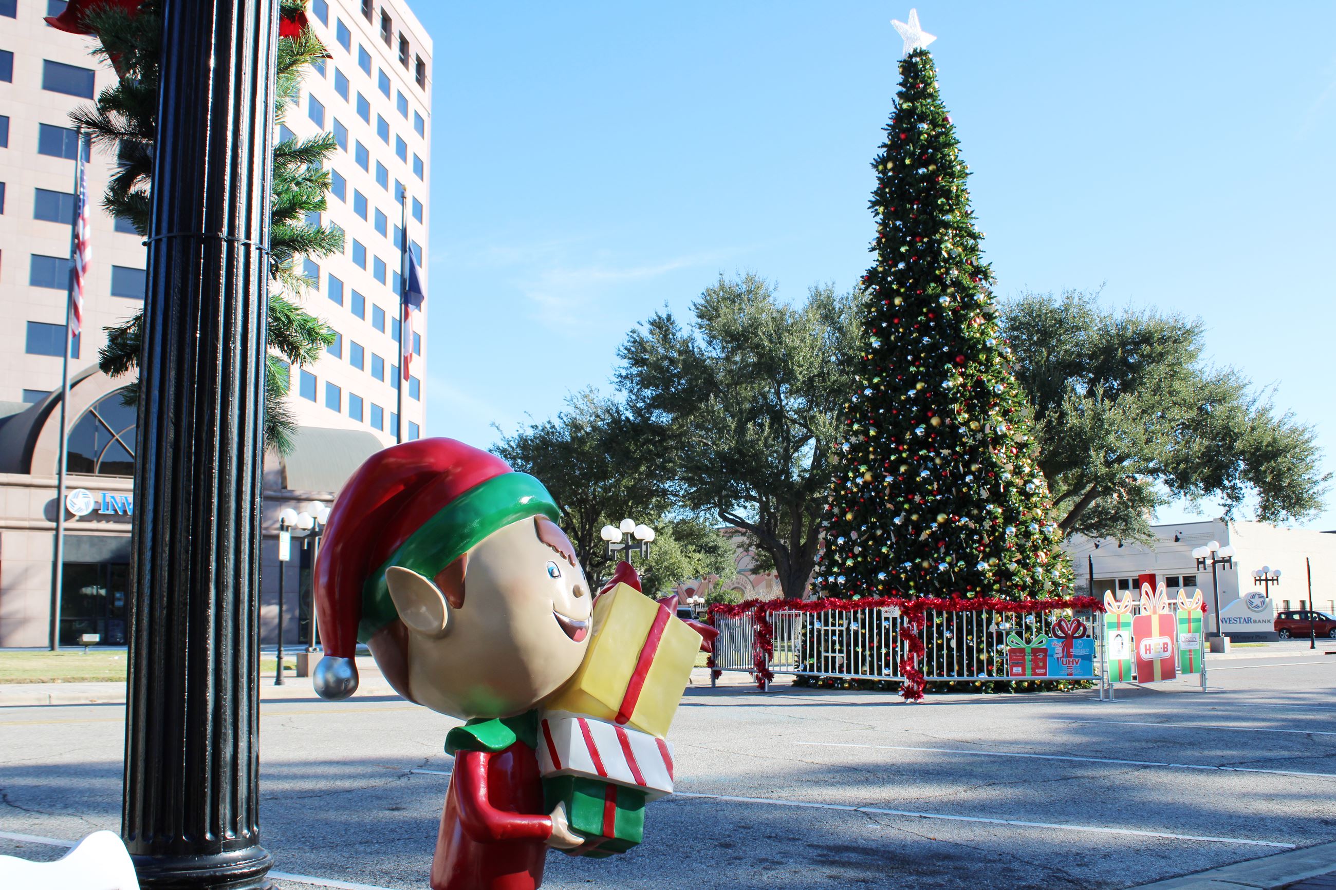 A plexiglass elf sculpture stands near a lamppost with the H-E-B Christmas Tree in the background.