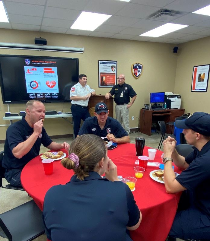 2 people stand at a podium in the background by a large presentation screen. 4 people eat at table.