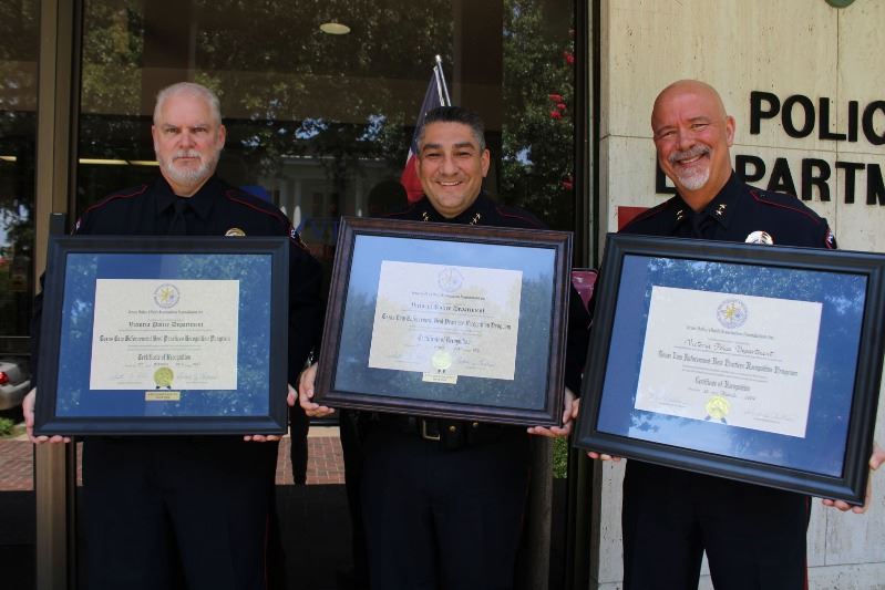 Three men in police uniform hold framed certificates outside police station