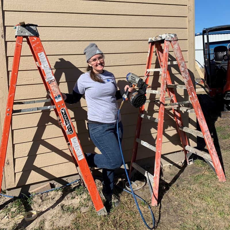 Woman leans on an exterior wall between two ladders holding a power tool