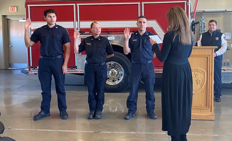 Three people stand with hands raised in front of a fire truck. A woman reads from a paper in front.