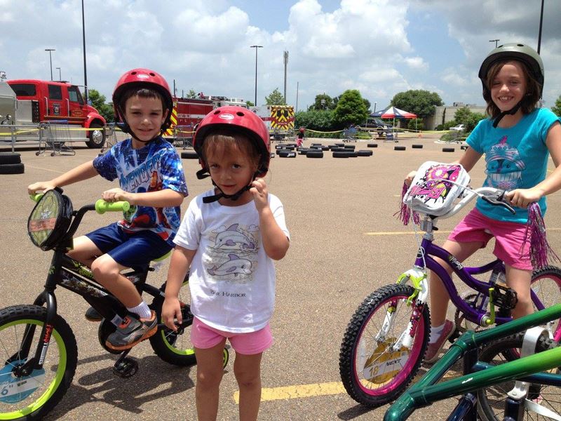 Three kids with bike helmets in a parking lot. Two are riding bikes.