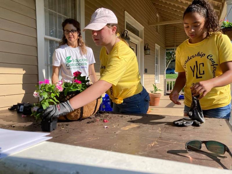 Two preteens in matching T-shirts arrange flowers in a planter; a woman in a KVB shirt supervises