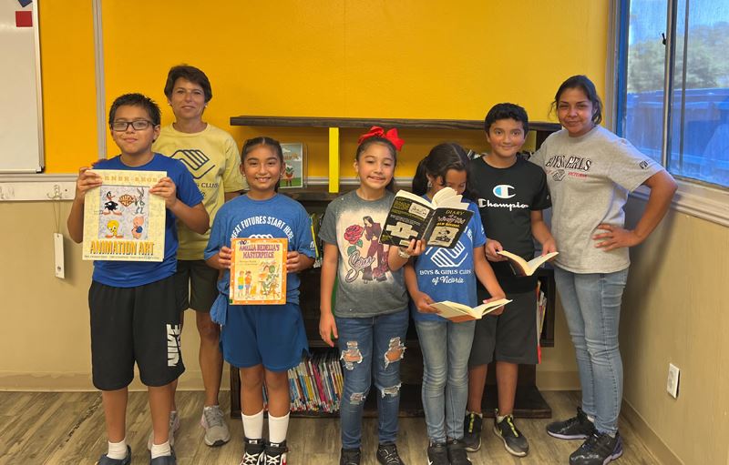 Kids and two adults pose with books in front of a bookshelf.