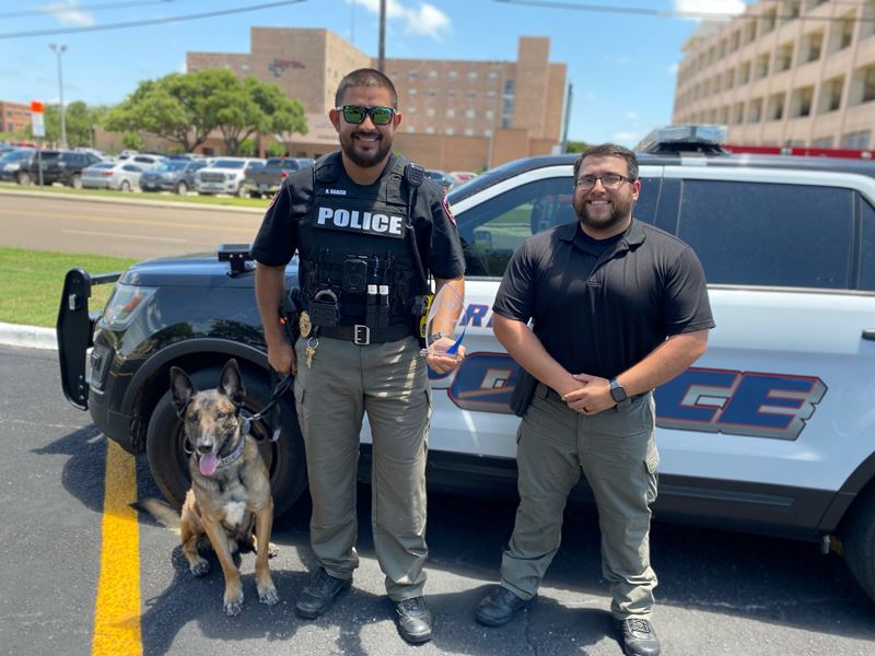 Two men stand near a police car with a dog on a leash
