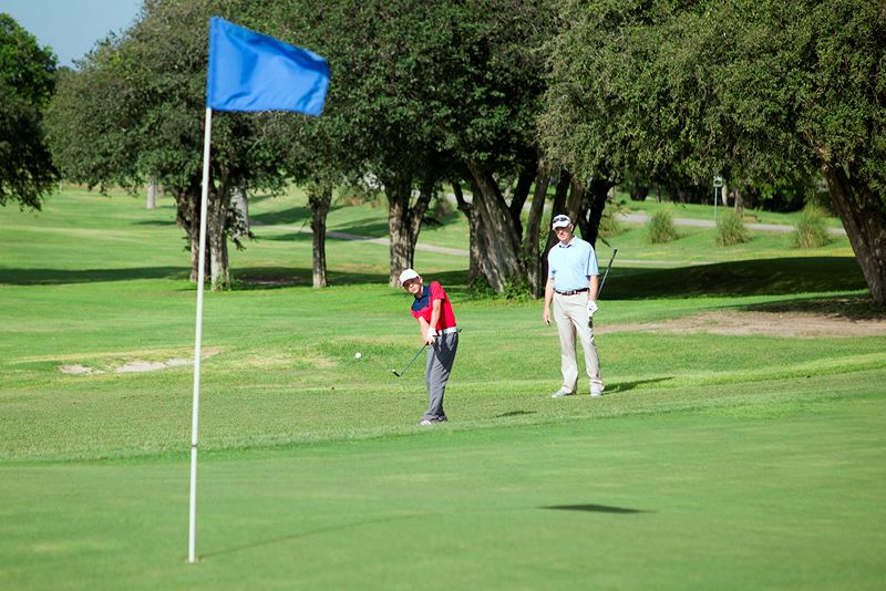 A young golf player hits a golf ball toward a hole while an adult watches