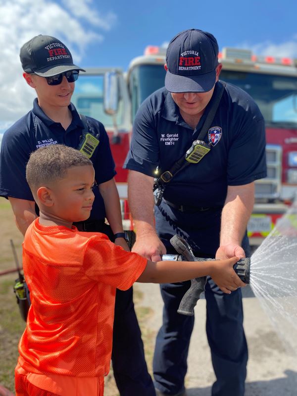 Two people in Victoria Fire Department caps and polos help a child aim a fire hose