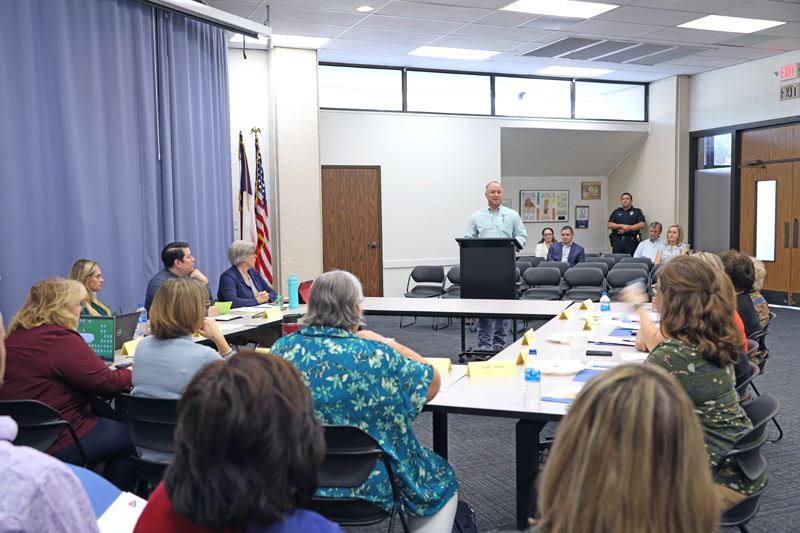 In a small meeting room, a man at a podium addresses many people seated at tables.