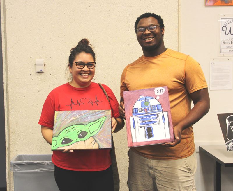 Two smiling people hold paintings of Star Wars characters Baby Yoda and R2D2.