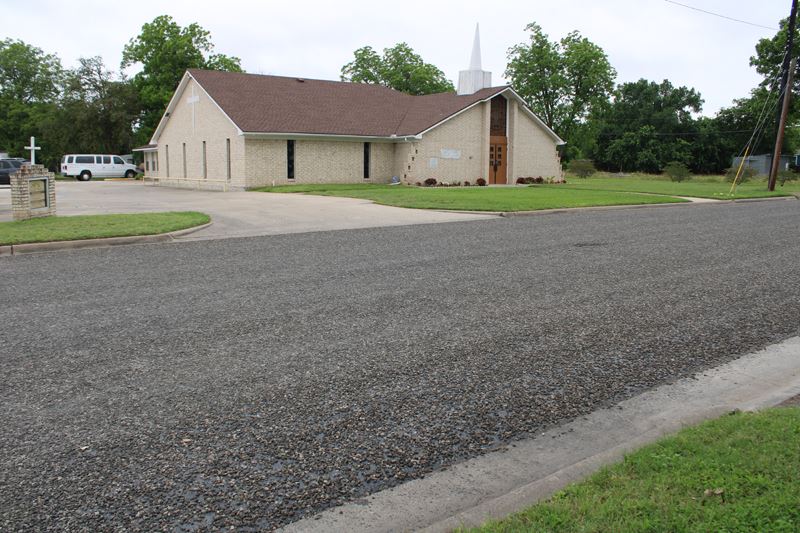 A roadway is coated in dark, rough gravel in front of a church.