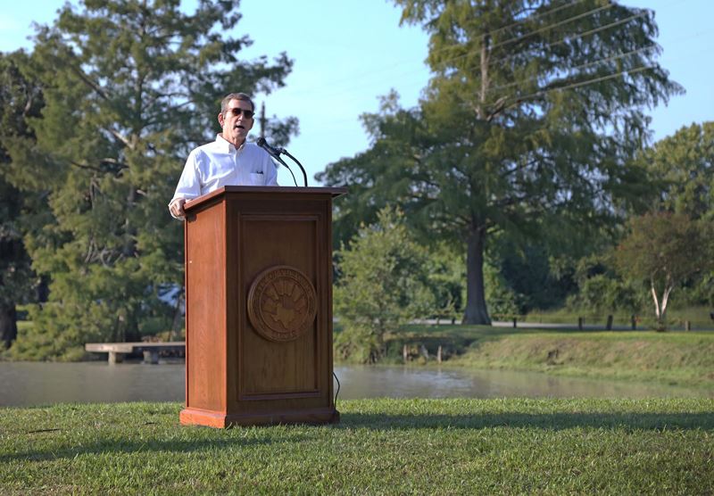 Man stands at podium with City seal in Riverside Park. There is a pond behind him.