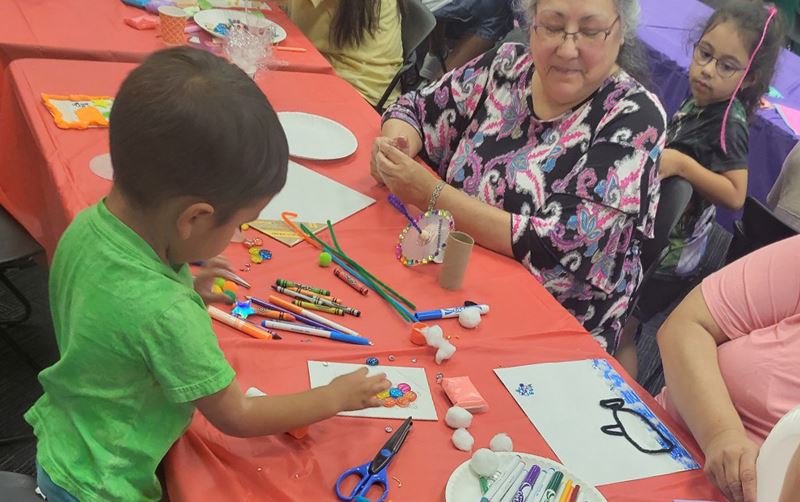 Adults and children sit at tables and make art using crayons, pipe cleaners and other supplies.