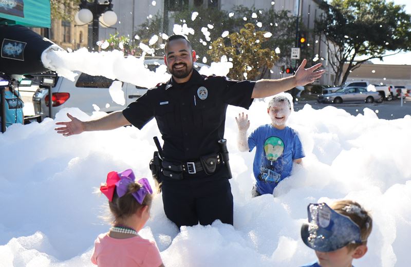 A uniformed police officer and three kids run around in foam bubbles