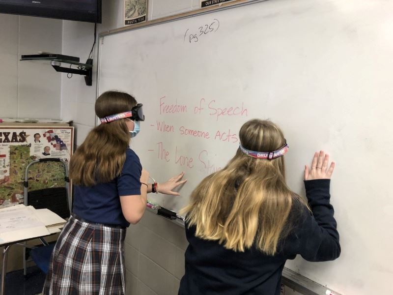 Two girls wearing impairment goggles stand close to a whiteboard with large lettering written on it.