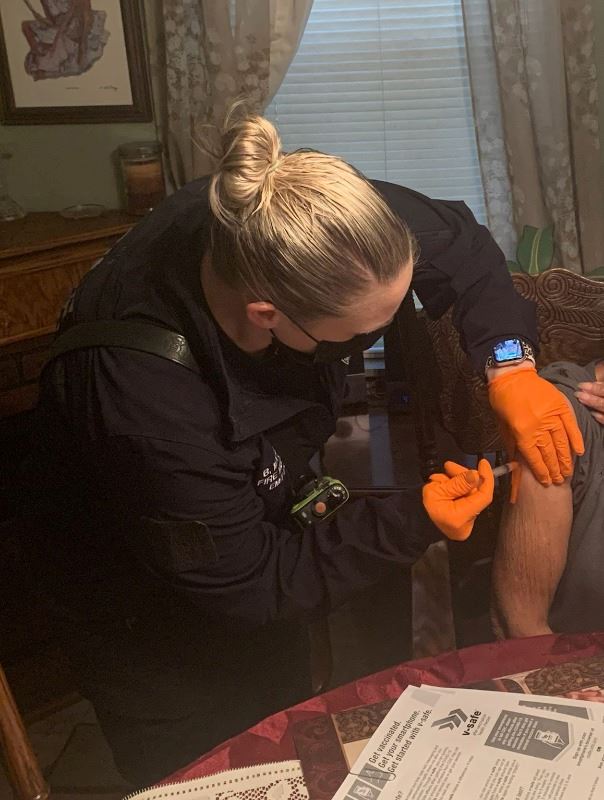 A woman wearing medical gloves uses a syringe to give an injection to a seated elderly woman