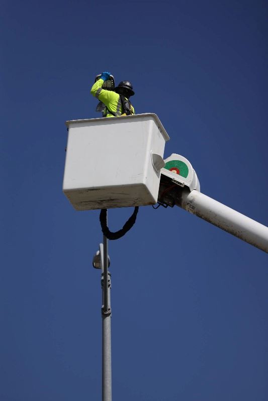 A worker in a yellow vest in the bucket of a cherry picker truck changes a bulb on a street light