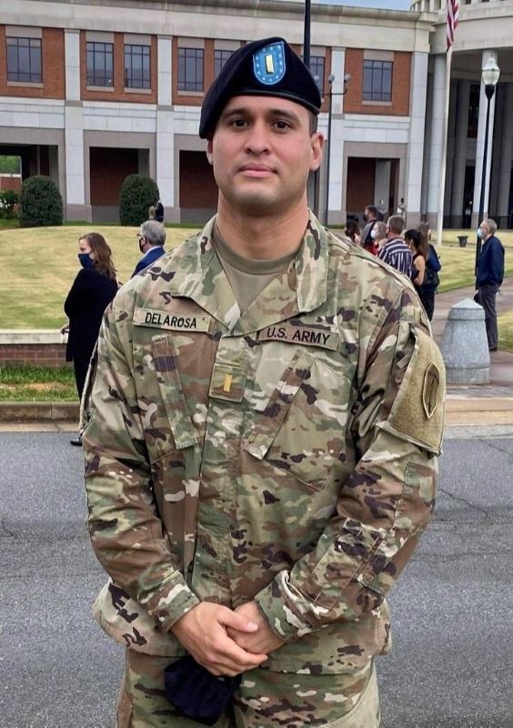 A man in a U.S. Army uniform stands outside with a large brick building and people in the background
