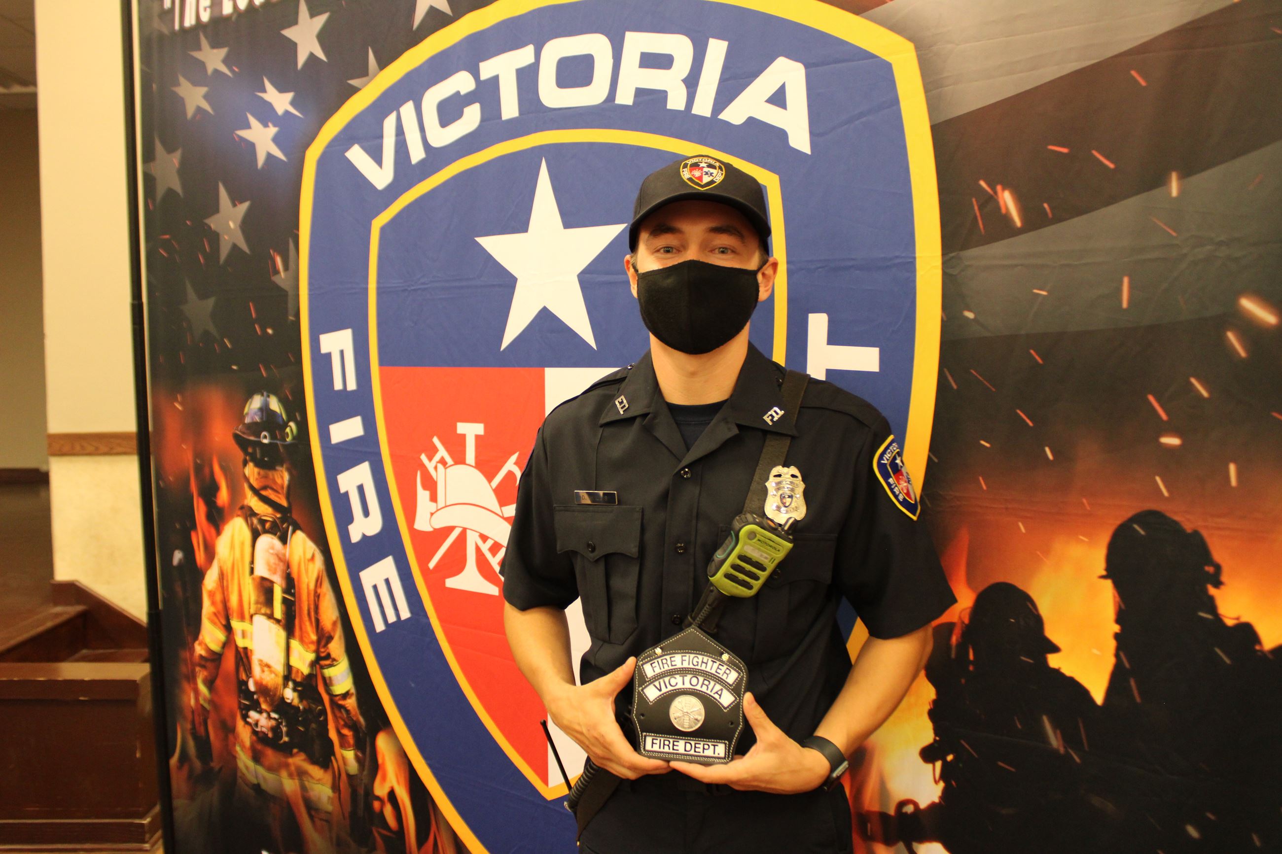 Young man holding firefighter patch stands in front of Victoria Fire Department backdrop.