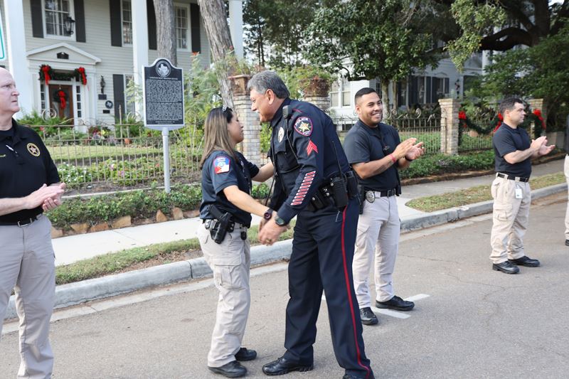 An older police officer hugs a younger officer who is standing in a row of applauding officers.