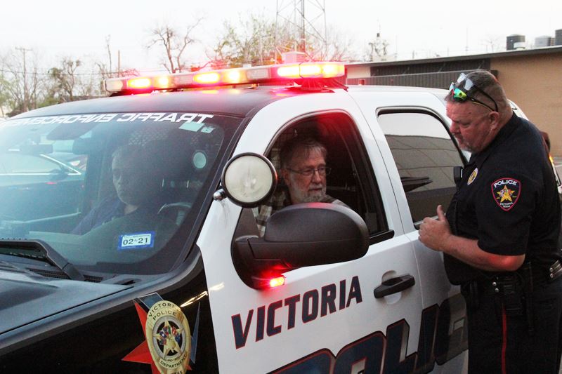 A uniformed police officer speaks to two people sitting in the front of a police car