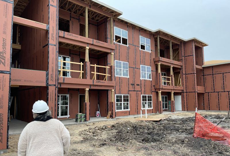 A woman in a hard hat stands outside an unfinished apartment building.