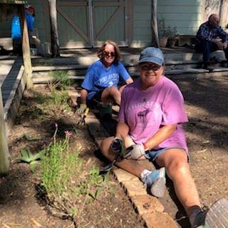 Two women sitting next to a narrow flower bed holding new plants they are planting there